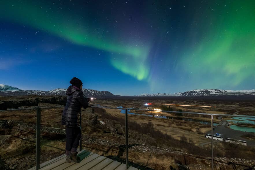 Person watching green northern lights above Thingvellir National Park, with snow-capped mountains and winding rivers in the Icelandic landscape below.