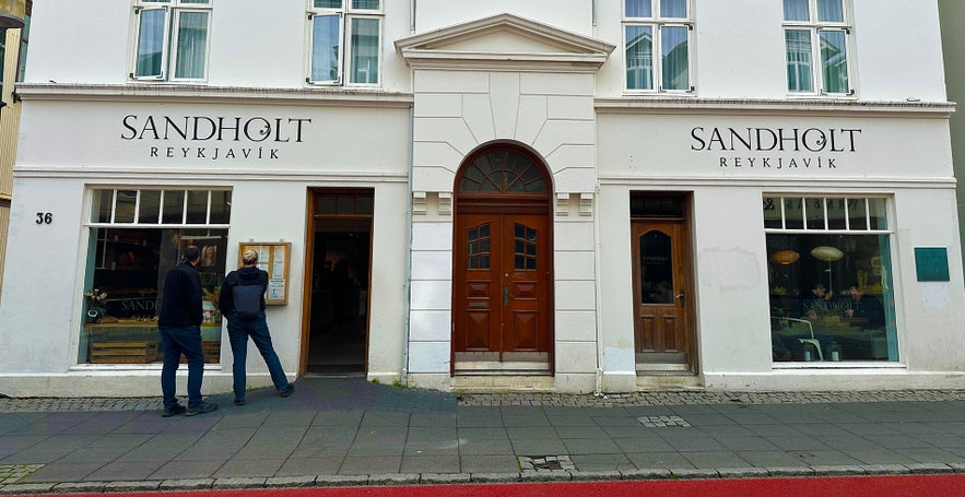 Street view of Sandholt Bakery in Reykjavik with white facade and visitors reading the menu.