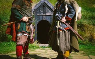 Two Viking reenactors in traditional clothing stand outside a turf house, holding an axe and a sword in a grassy Icelandic setting.