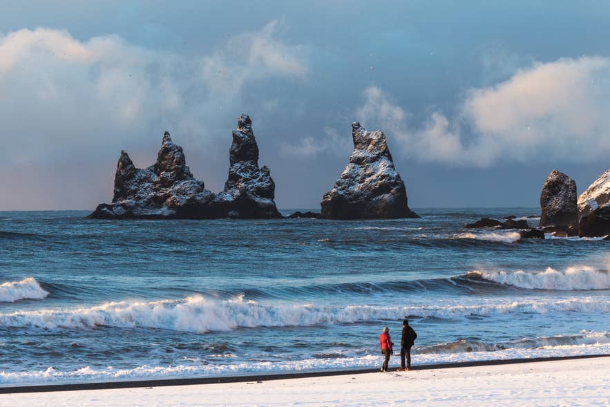 Two travelers standing on a black sand beach facing Reynisdrangar sea stacks near Vik, Iceland