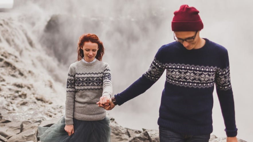 A couple wearing replica lopapeysa sweaters walking hand in hand near a misty Icelandic waterfall in a cozy outdoor scene.