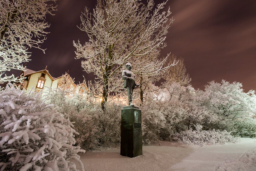 Statue of poet Jónas Hallgrímsson surrounded by snow-covered trees and bushes in Reykjavík's Austurvöllur park on a winter night.