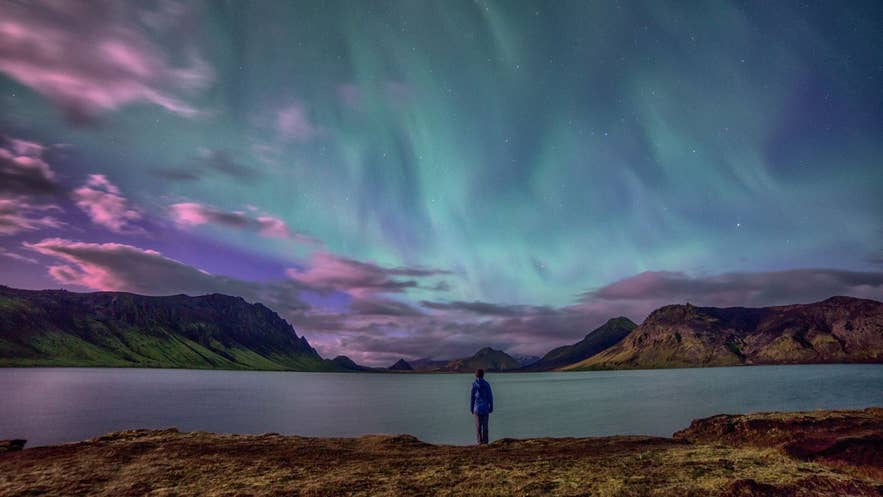 Aurores boréales au-dessus d’un lac en Islande, avec une personne observant l’aurore et un ciel coloré se reflétant sur les montagnes la nuit.