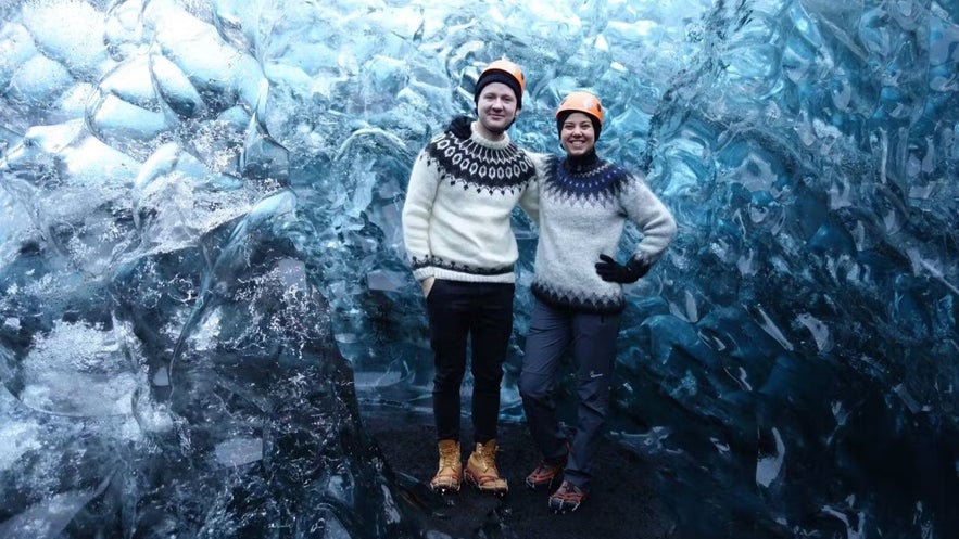 Couple in lopapeysa sweaters standing inside a glacial ice cave, dressed warmly for a winter tour in Iceland.