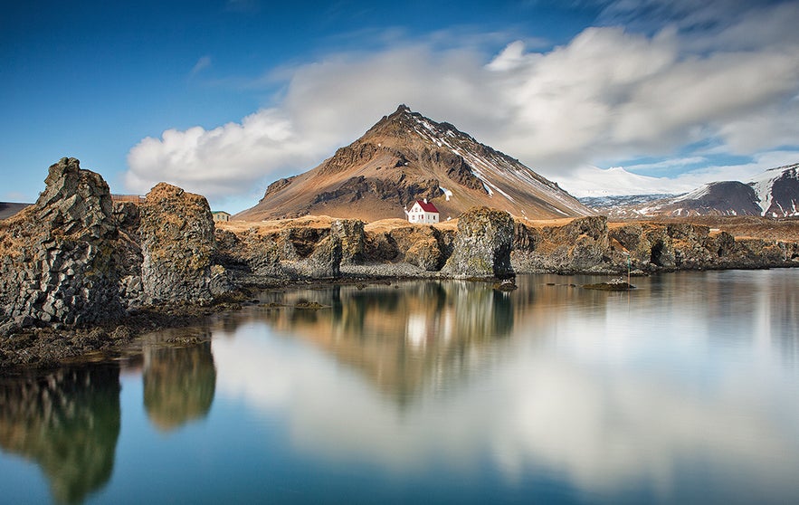 Arnarstapi fishing village on the Snæfellsnes Peninsula in West Iceland, with coastal lava formations and a white house beneath a snow-capped mountain.