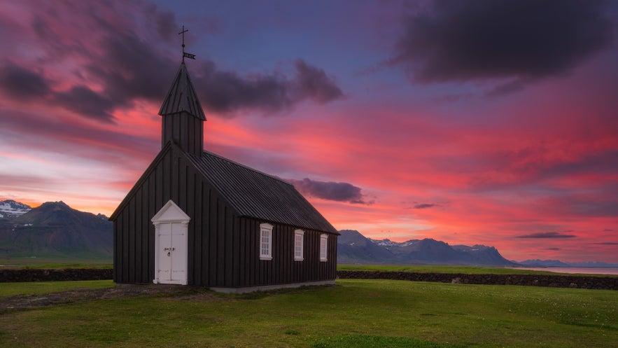 Búðakirkja black church on the Snæfellsnes Peninsula, Iceland, at sunset with dramatic pink and purple skies and distant mountains in the background.