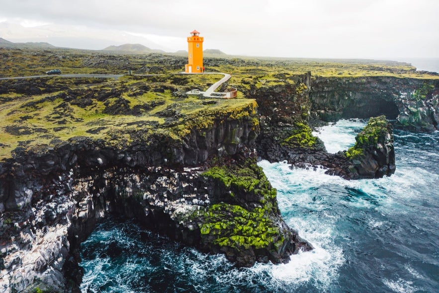Bright orange Svörtuloft lighthouse perched on mossy black lava cliffs above the Atlantic Ocean on Iceland’s Snæfellsnes Peninsula during overcast weather.