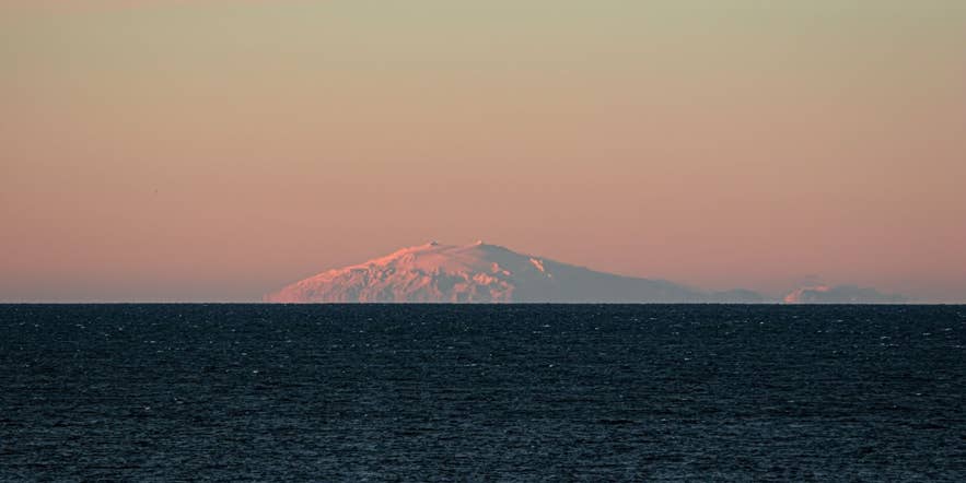 Snæfellsjökull glacier glowing in pink hues at sunset, viewed across the ocean from Iceland’s west coast on a clear, serene evening.
