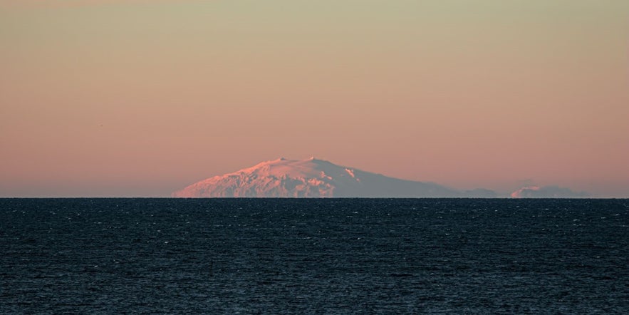Snæfellsjökull glacier glowing in pink hues at sunset, viewed across the ocean from Iceland’s west coast on a clear, serene evening.