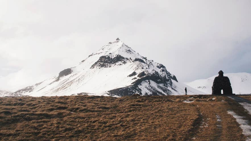 Snow-covered mountain behind the Bárður Snæfellsás statue in Arnarstapi, Snæfellsnes Peninsula, Iceland, with a lone figure walking on a winter path.