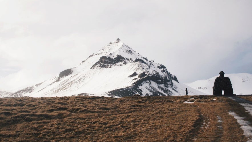 Snow-covered mountain behind the Bárður Snæfellsás statue in Arnarstapi, Snæfellsnes Peninsula, Iceland, with a lone figure walking on a winter path.