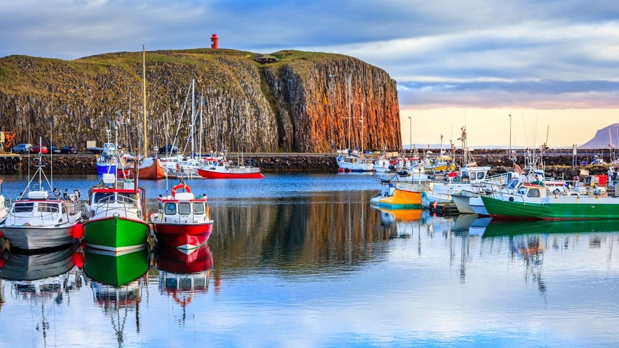 Colorful fishing boats docked in Stykkishólmur harbor with basalt cliff and red lighthouse in the background, Snæfellsnes Peninsula, Iceland.