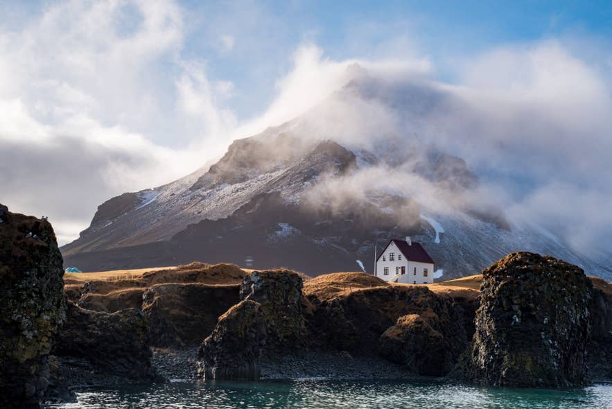 Zu sehen sind Küstenklippen und ein weißes Haus unter einem nebelverhangenen Berg in Arnarstapi auf der Snaefellsnes-Halbinsel.