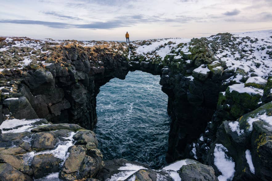 Ein Wanderer steht auf dem natürlichen Felsbogen bei Arnarstapi auf der Snaefellsnes-Halbinsel im Winter.