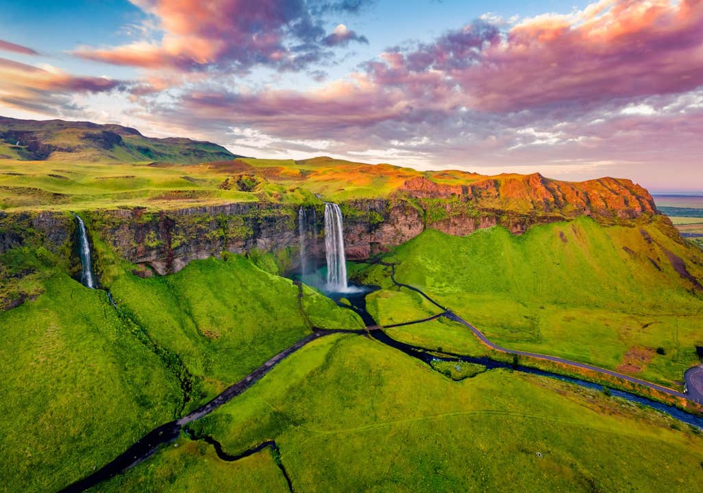 Aerial view of Seljalandsfoss waterfall cascading over a cliff into lush green fields at sunset, South Iceland.