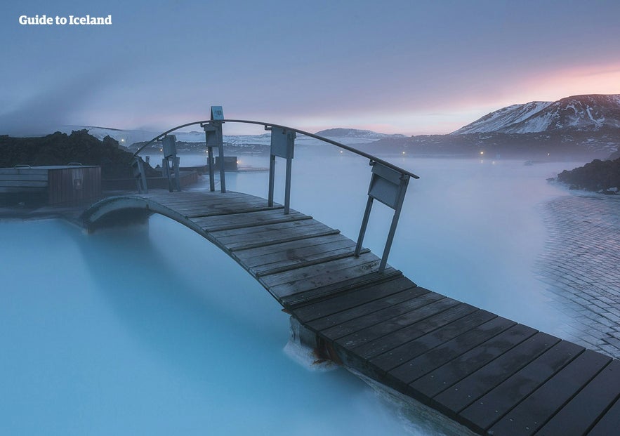 Wooden bridge over Blue Lagoon geothermal spa in Iceland at sunrise, with milky blue water and snow-covered volcanic landscape.