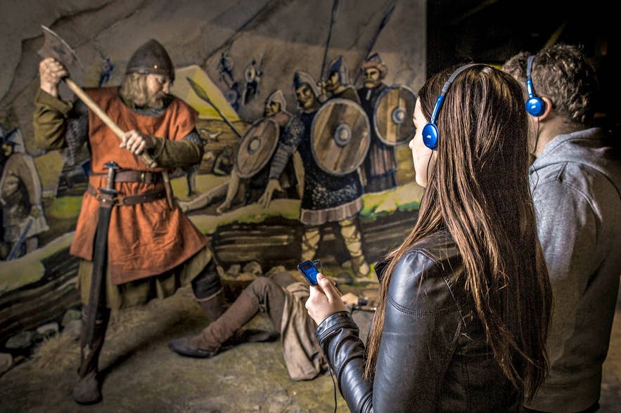 A museum exhibit depicting a Viking warrior raising an axe over a fallen figure, with two visitors wearing blue headphones observing the scene at the Saga Museum in Reykjavik.