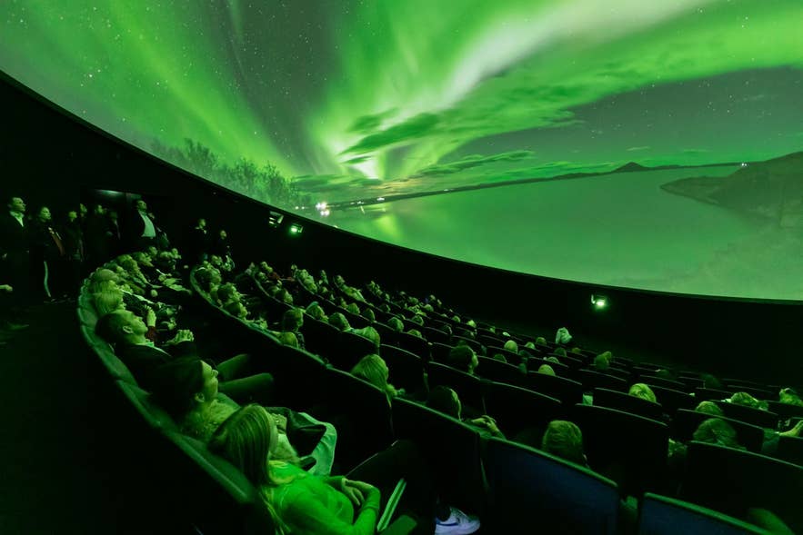 Bezoekers kijken naar de Áróra noorderlicht-planetariumshow in Museum Perlan in Reykjavik, IJsland, met levendige groene aurora-beelden geprojecteerd op het koepelscherm.