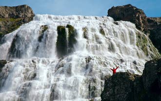 A person stands on a superb viewing rock in front of Dynjandi waterfall with their arms outstretched and the water cascading powerfully behind them.