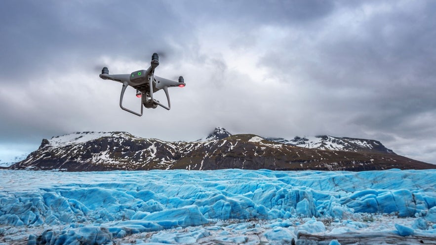 A drone flying over bright blue glacial ice with snow-capped mountains in the background under a cloudy sky in Iceland.