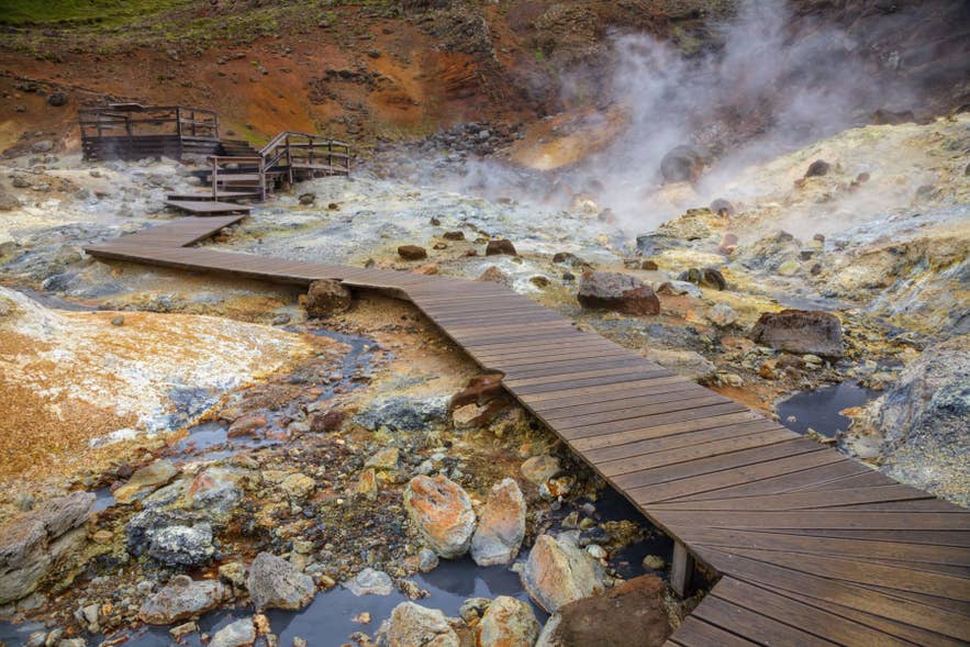 Holzsteg, der sich durch ein dampfendes Geothermalgebiet in Island mit blubbernden heißen Quellen und mineralisch verfärbtem Boden schlängelt. Holzsteg, der sich durch ein dampfendes Geothermalgebiet in Island mit blubbernden heißen Quellen und mineralisch verfärbtem Boden schlängelt.