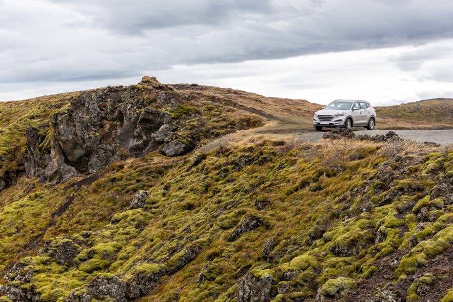 Rental car driving on a narrow mountain road through mossy lava fields in rural Iceland.