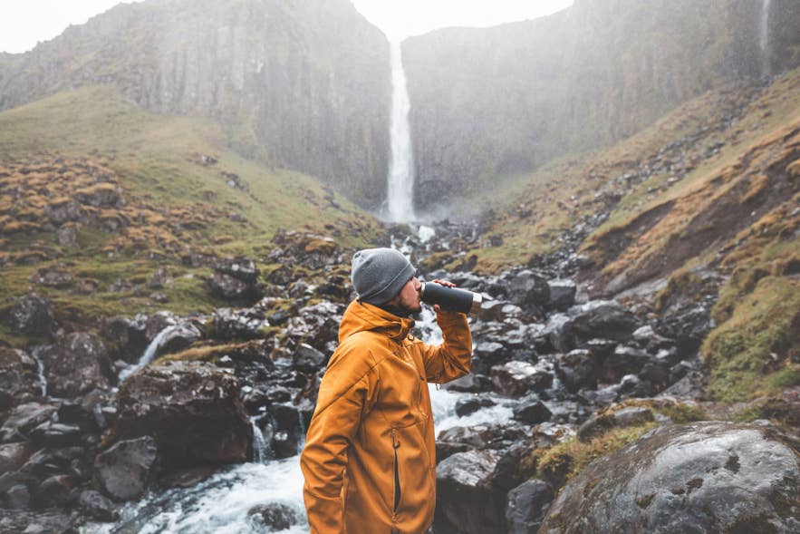 Traveler drinking from reusable bottle near a waterfall in Iceland, highlighting Iceland’s clean tap water.