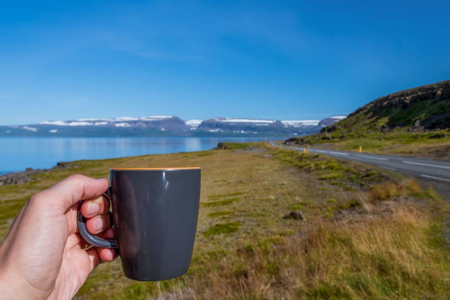 Hand holding coffee mug along Route 1 Ring Road with Icelandic fjord landscape in Westfjords, Iceland. Hand holding coffee mug along Route 1 Ring Road with Icelandic fjord landscape in Westfjords, Iceland.