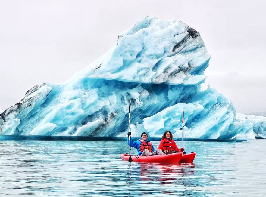 Kajakfahren in der Gletscherlagune Jökulsarlon im Juni in Island, zwei Paddler in roter Ausrüstung gleiten an einem blauen Eisberg vorbei.