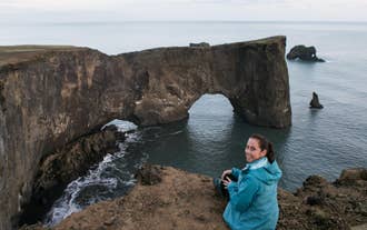 A woman poses for a photo on a cliff facing the Dyrholaey Rock in Iceland.