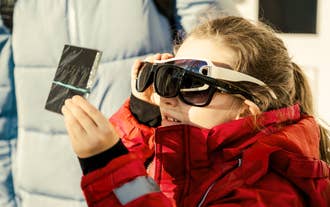 A child wears a pair of eclipse glasses and uses a filter to view the eclipse.