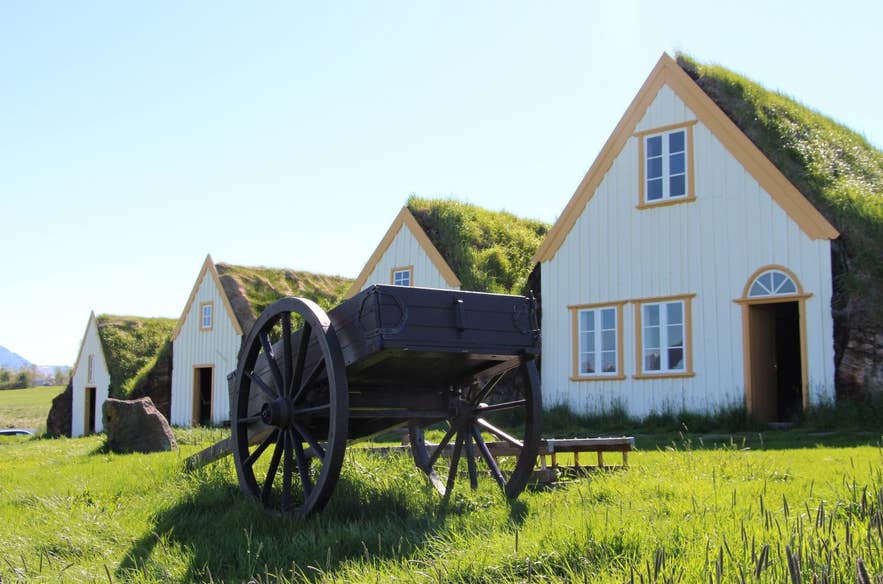 Historic turf-roofed farmhouses at the Glaumbaer Farm and Museum in northern Iceland, with a wooden cart in the grassy foreground.