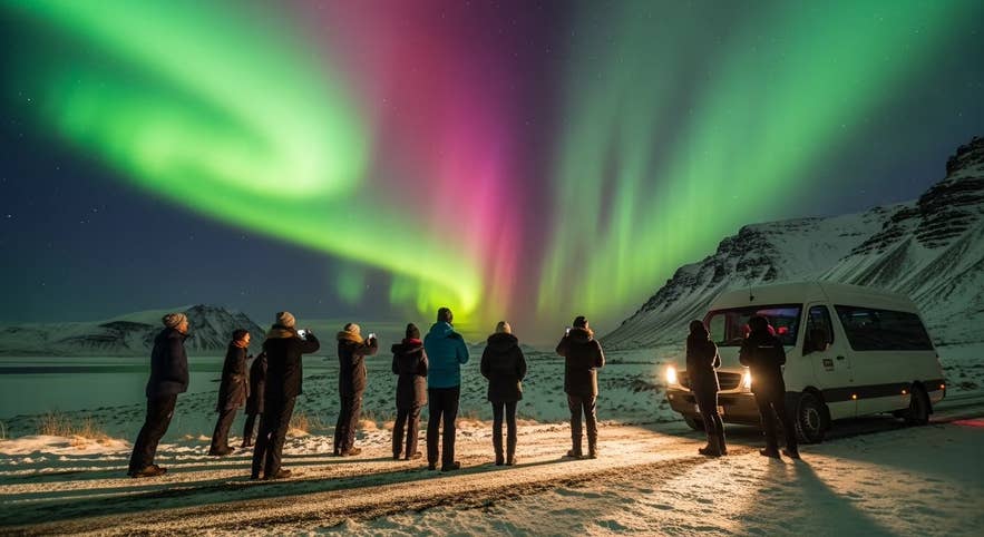 Reisende auf einer mehrtägigen geführten Wintertour durch Island halten an, um neben ihrem Reisebus die leuchtenden Nordlichter über den schneebedeckten Bergen zu fotografieren.
