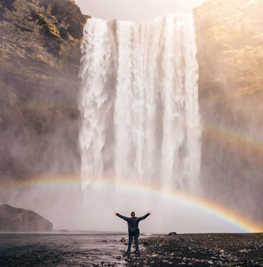 Een persoon staat voor de machtige Skogafoss-waterval aan de zuidkust van IJsland met zijn armen wijd uitgestrekt terwijl een regenboog de voorkant van de waterval siert.
