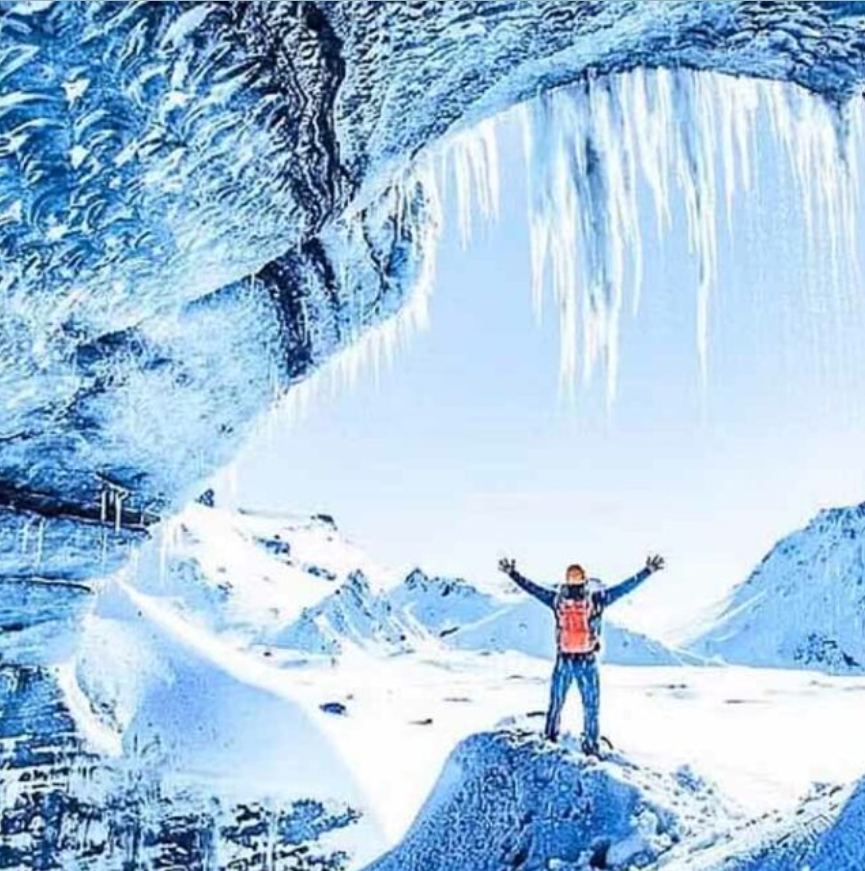 Een persoon staat in de ingang van een ijsgrot in IJsland met zijn armen uitgestrekt en wordt omringd door een spectaculair landschap.