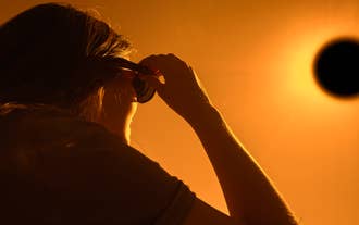 A person watches a solar eclipse using protective glasses against a bright orange sky.