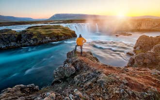 Traveler standing near Godafoss Waterfall in North Iceland at sunset with flowing water and golden light.