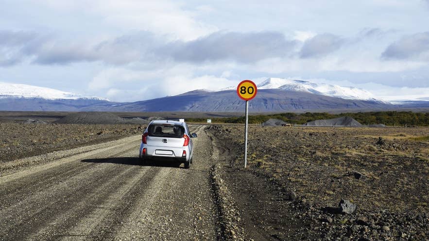 A small rental car parked on an Icelandic gravel road beside a road sign, with a glacier in the distance.