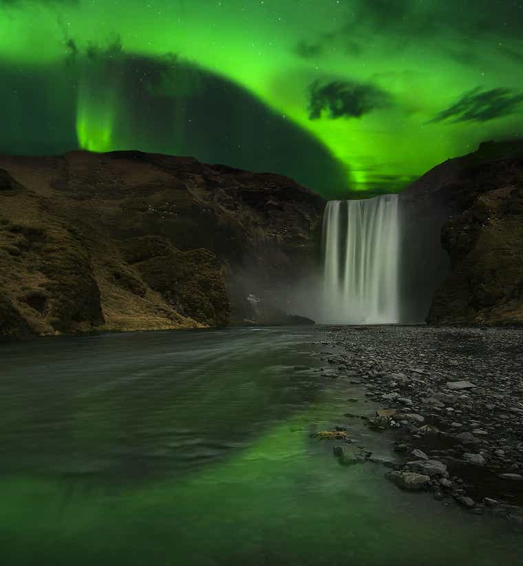Northern lights over Skogafoss Waterfall at night, with green reflections on the river and dark cliffs in the background.