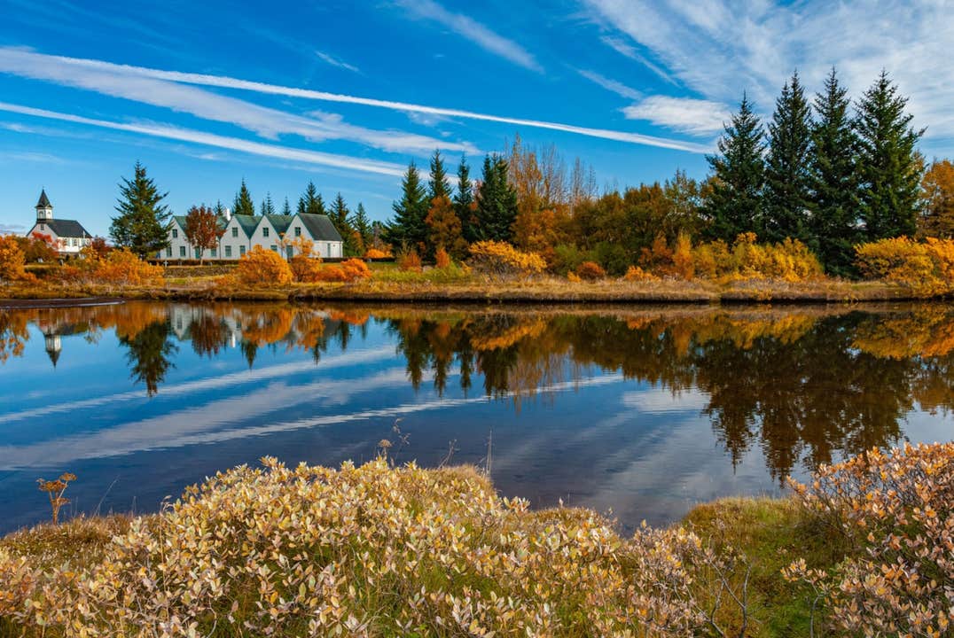 Thingvellir National Park in Iceland with autumn foliage reflected in calm water, historic buildings, and trees under a blue sky