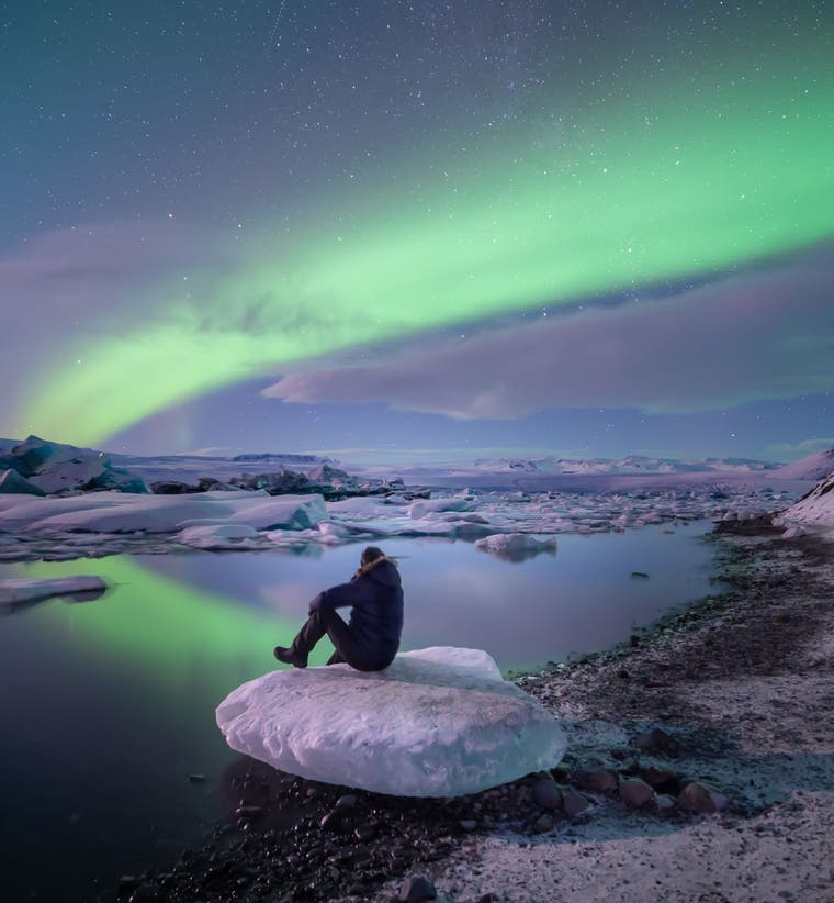 A solo traveler sits on ice at Jokulsarlon Glacier Lagoon in Iceland under the northern lights reflecting on the water.