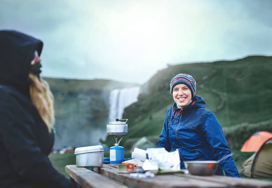 Travelers cooking a simple meal at a campsite in Iceland near a waterfall during a road trip camping stop.