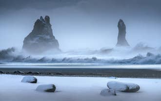 Waves crash on Reynisfjara Beach in South Iceland as black sand and sea stacks rise under a cloudy sky.