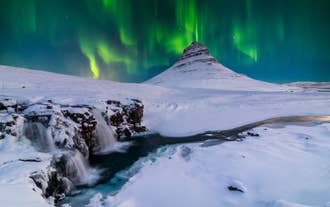 Kirkjufell Mountain stands in West Iceland as northern lights dance above the iconic peak and nearby waterfall at night.