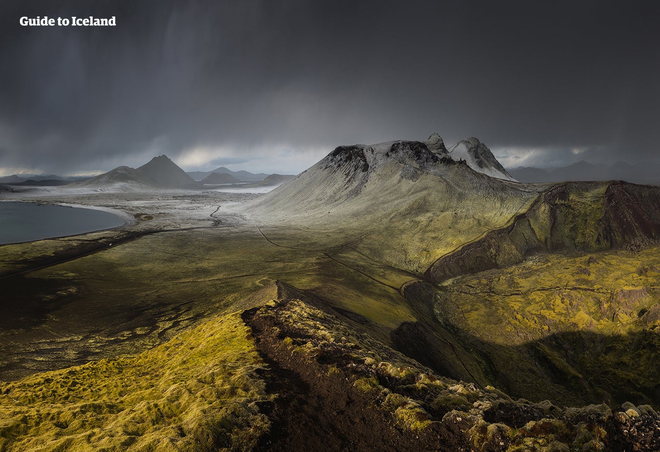 A light dusting of snow and a dark, moody sky present a striking contrast against a green mountain range in the Icelandic Highlands.