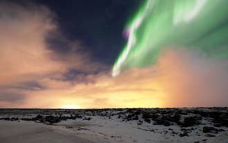 A dash of green and white from the aurora borealis contrasts beautifully with the dark sky and colors from the setting sun above a snowy landscape.