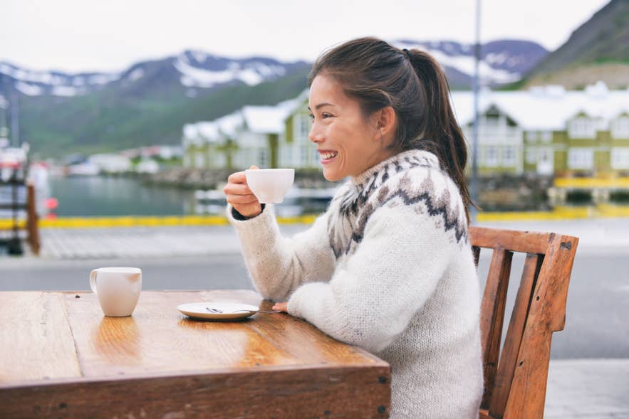 Woman wearing Icelandic wool sweater drinking coffee by scenic harbor in Iceland.