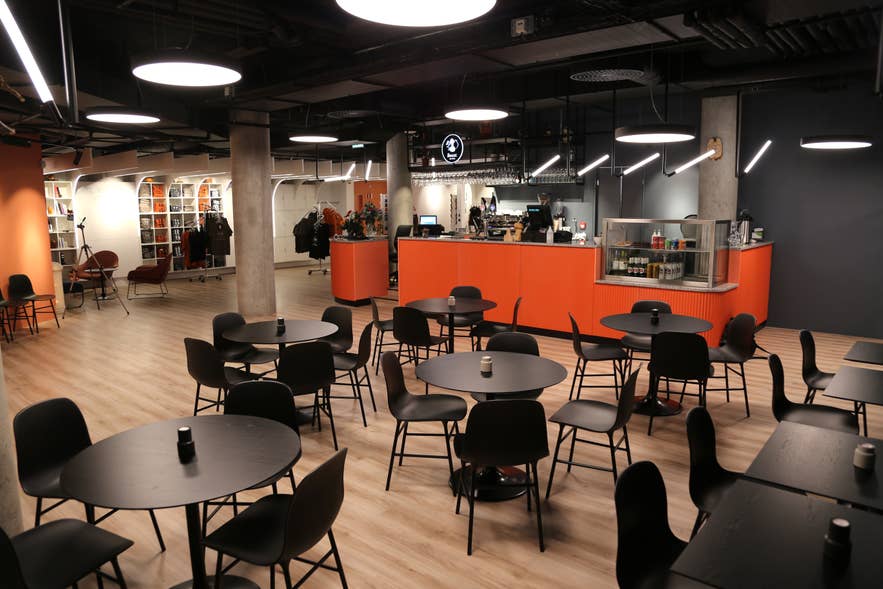 Interior of The Phallic Cafe at the Icelandic Phallological Museum, featuring modern black tables, chairs, pendant lighting, and an orange counter with shelves of merchandise in the background.