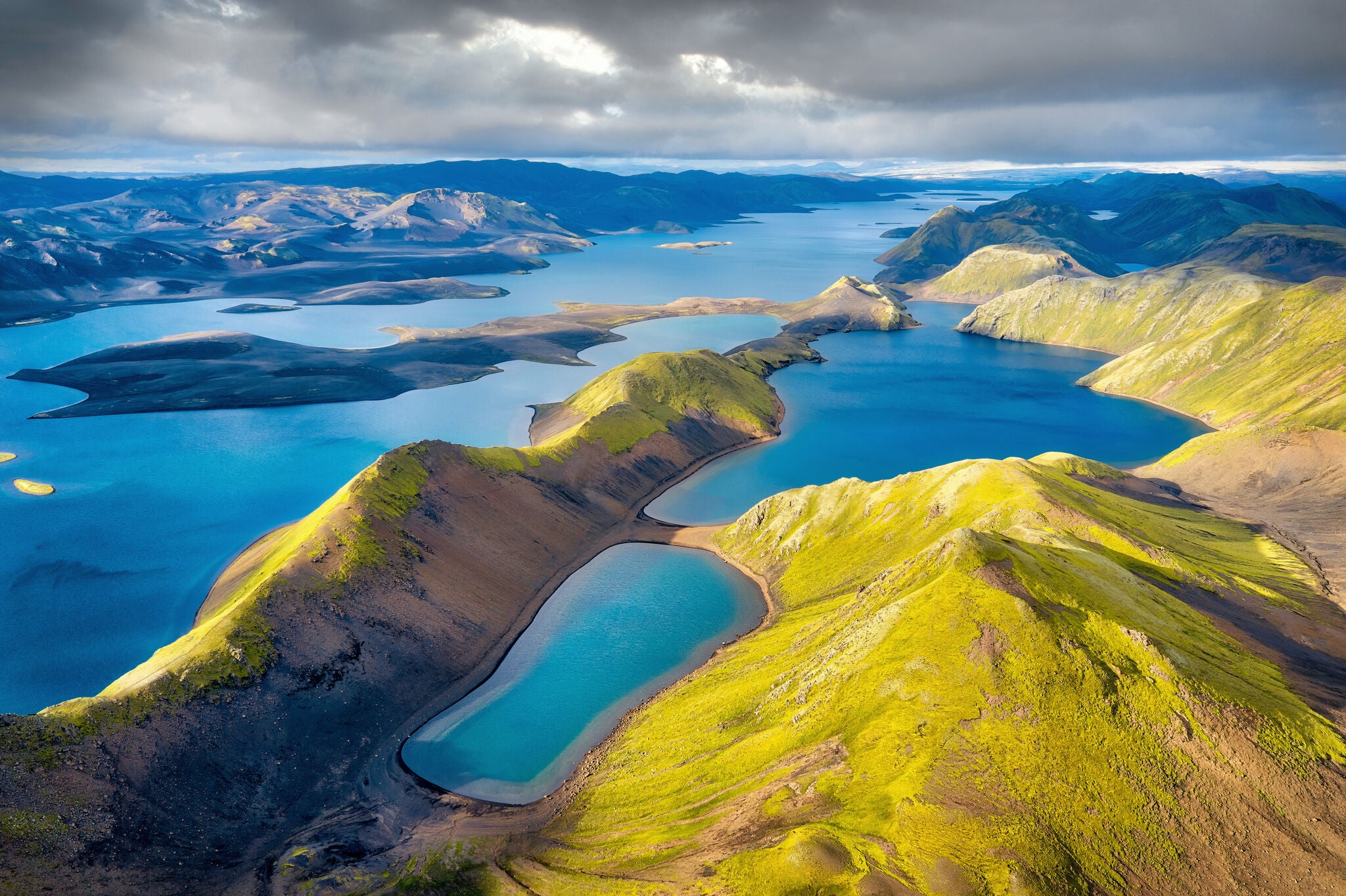Vue aérienne du lac Langisjor dans les Hautes Terres d’Islande, avec une eau d’un bleu intense serpentant entre des crêtes verdoyantes et volcaniques sous un ciel nuageux spectaculaire.