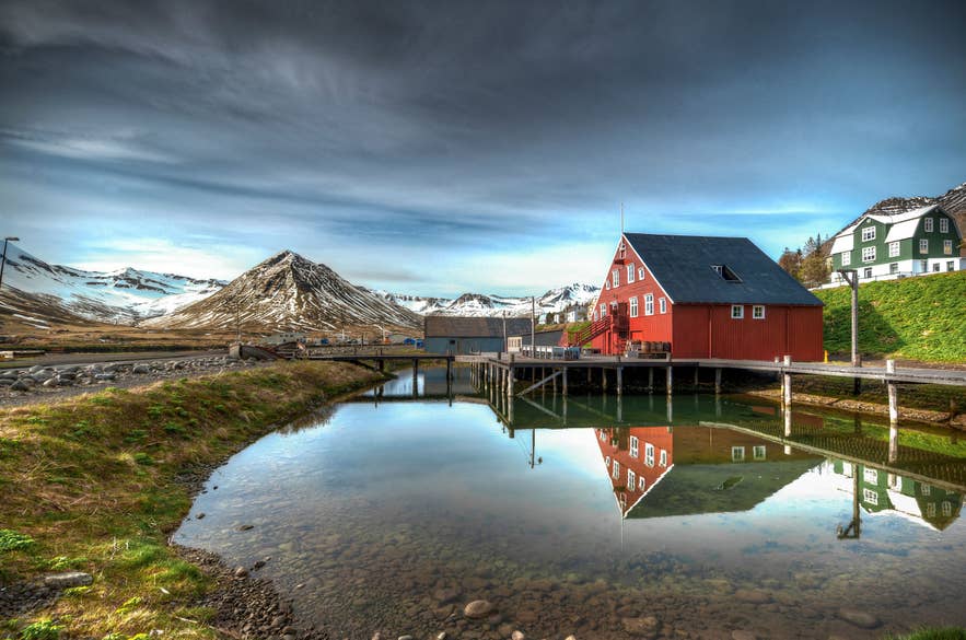 The Herring Era Museum in Siglufjordur fjord, featuring a red wooden building reflected in the calm harbor water.